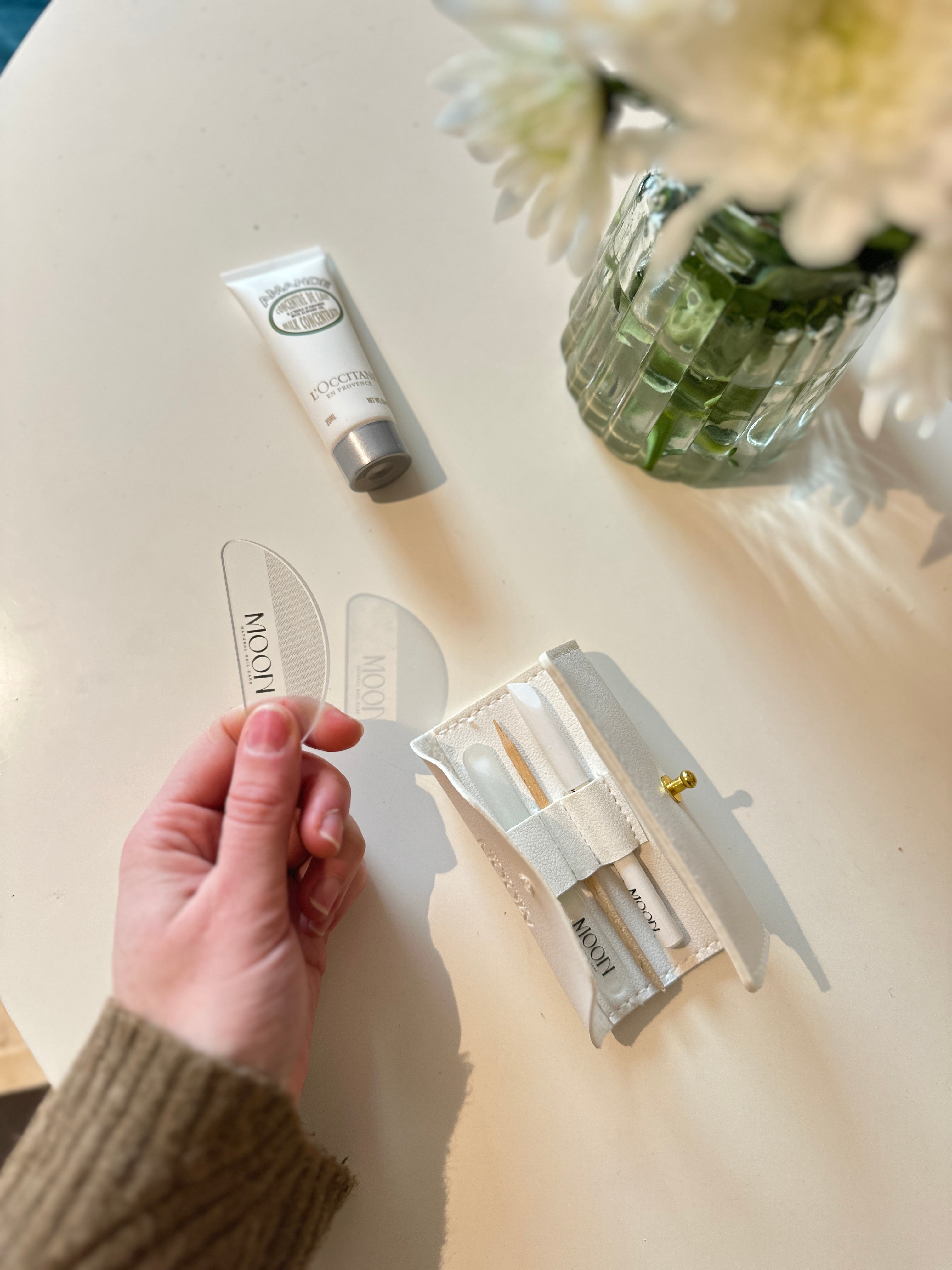 Hand holding a half-moon nail buffer with a tube and packaging on a light surface.